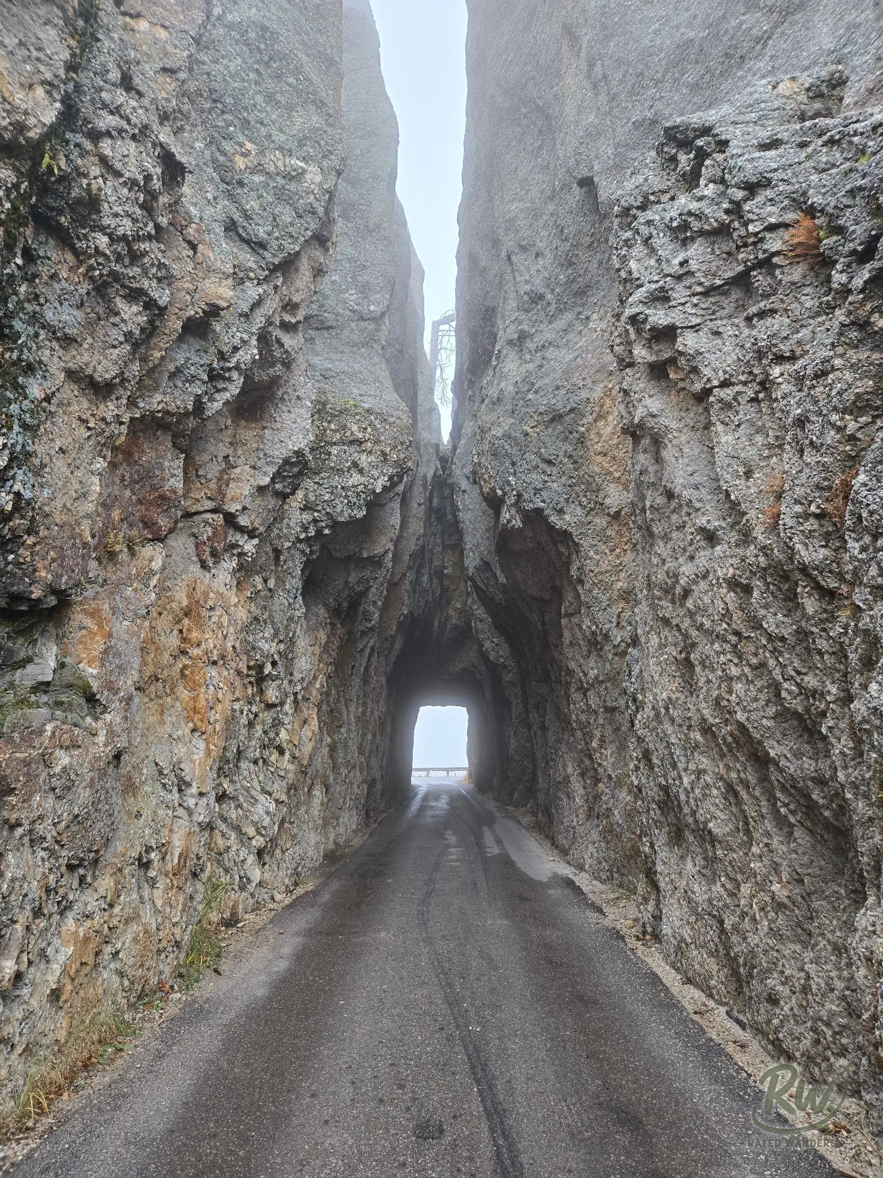 A narrow road running through a one-lane tunnel cut into a tall rock wall, fog above