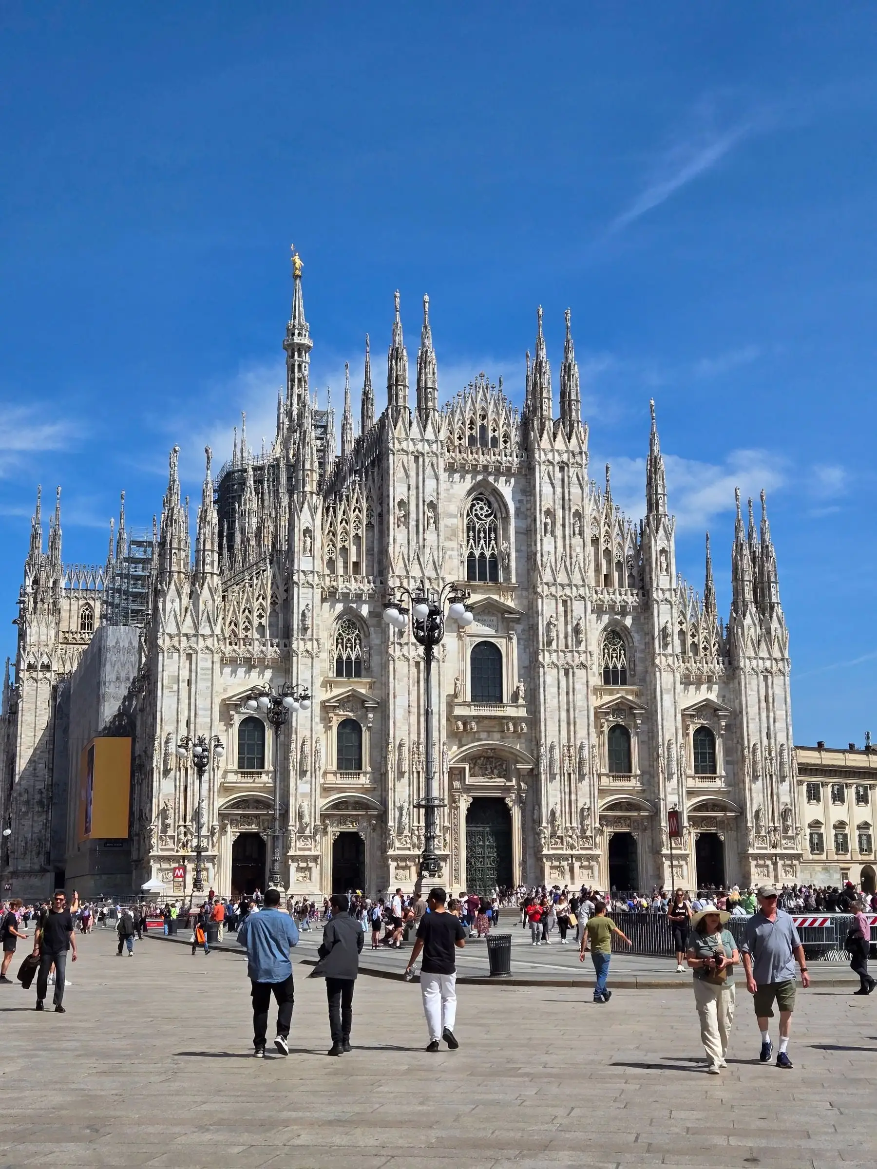 The white marble facade and spires of the Milan Duomo under a blue sky, visitors crossing the piazza
