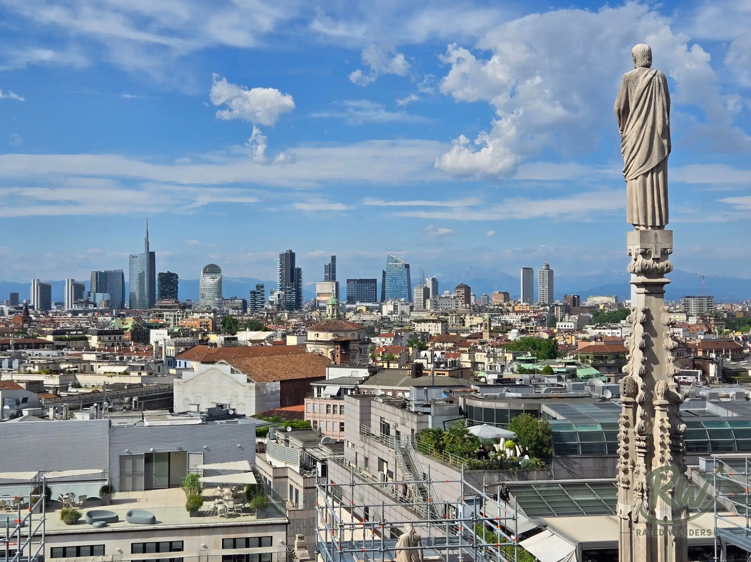 Milan’s rooftops and modern skyline seen from the Duomo terrace beside a carved statue