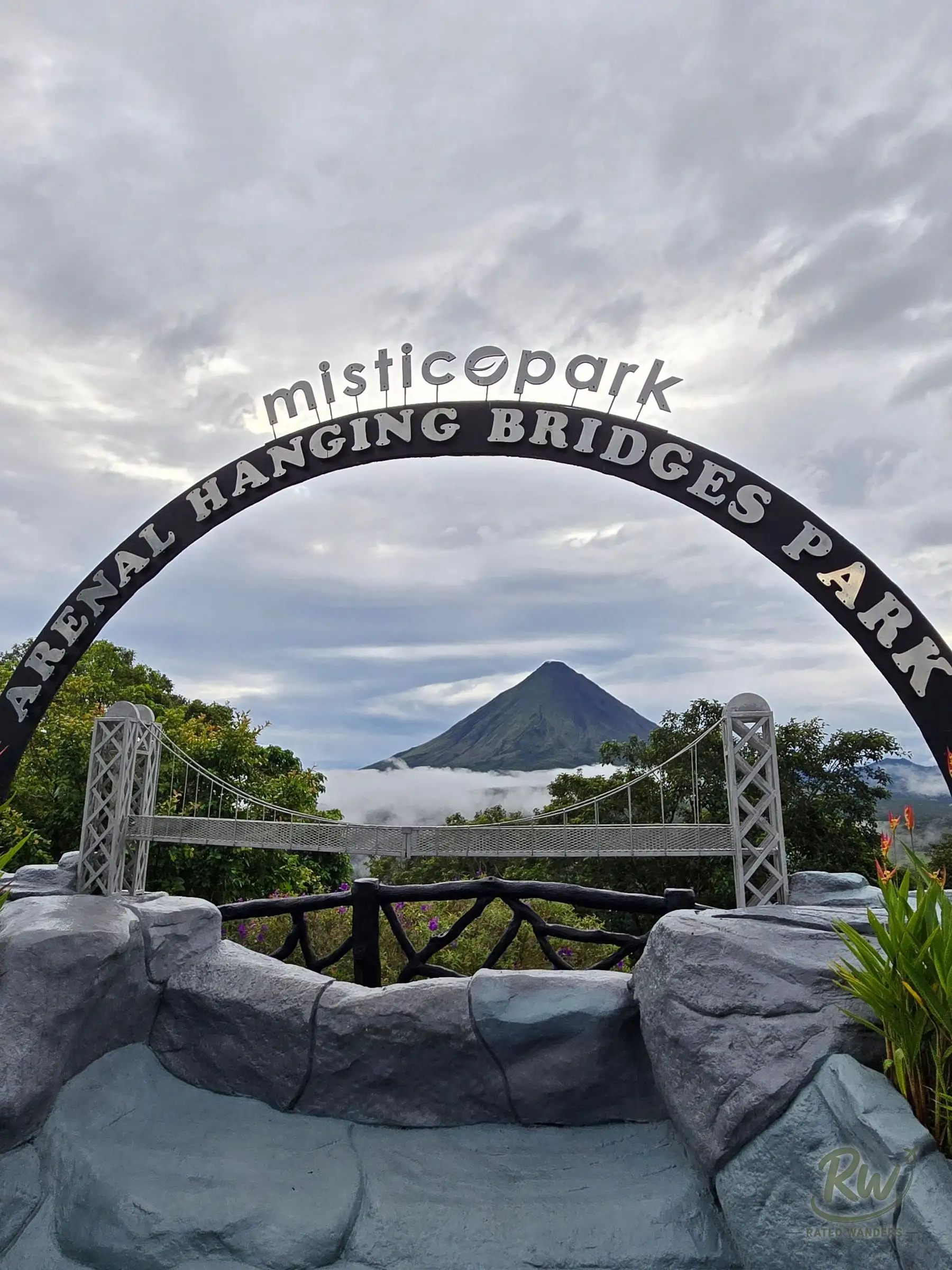 A miniature suspension bridge under the Mistico Park entrance arch, with Arenal volcano framed behind it