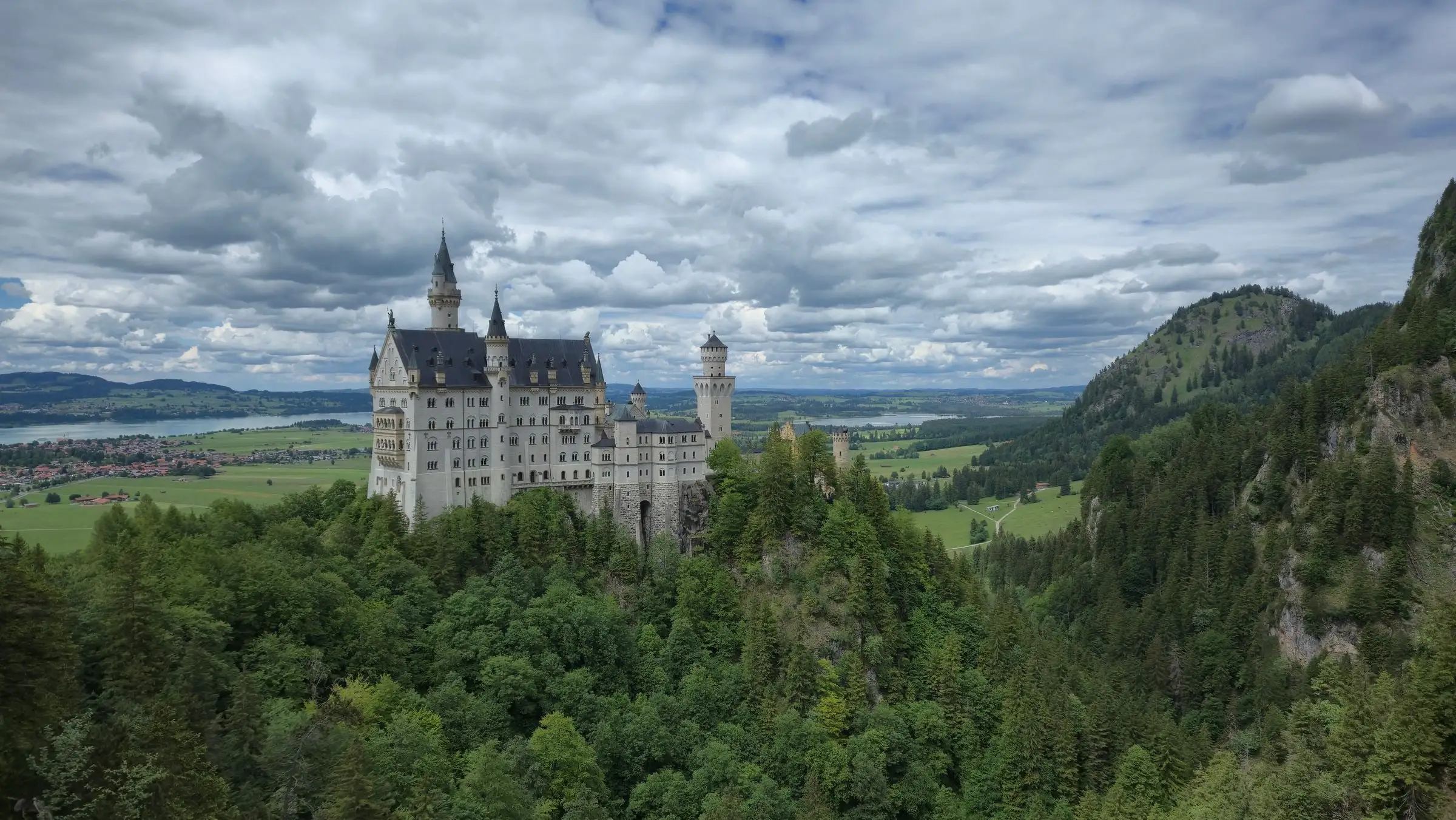 Neuschwanstein Castle on its wooded ridge under heavy cloud, with the Bavarian plain and a lake behind it
