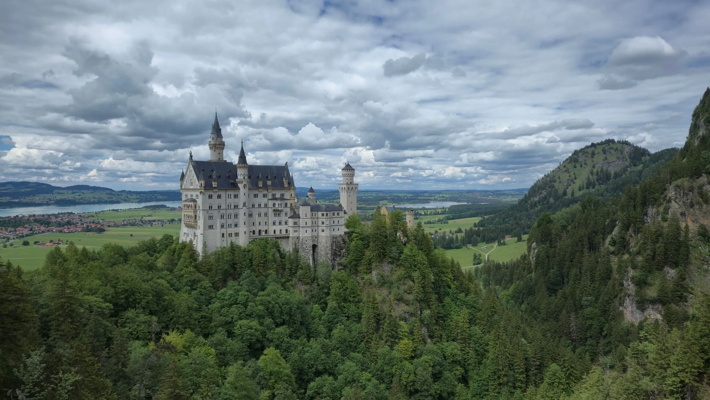Neuschwanstein Castle on its wooded ridge under heavy cloud, with the Bavarian plain and a lake behind it