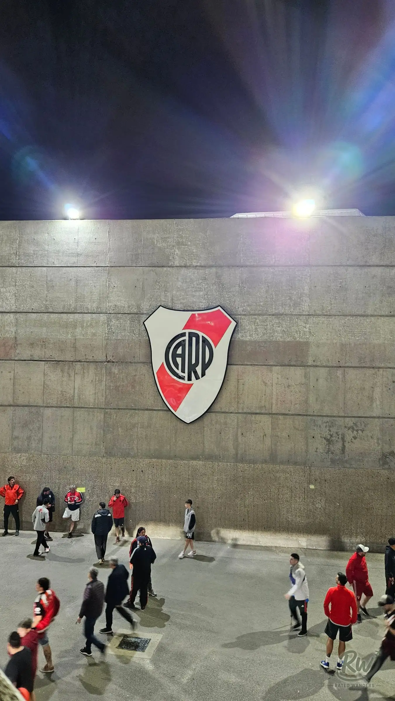 The River Plate crest on the concrete outer wall of the stadium, fans walking past under floodlights