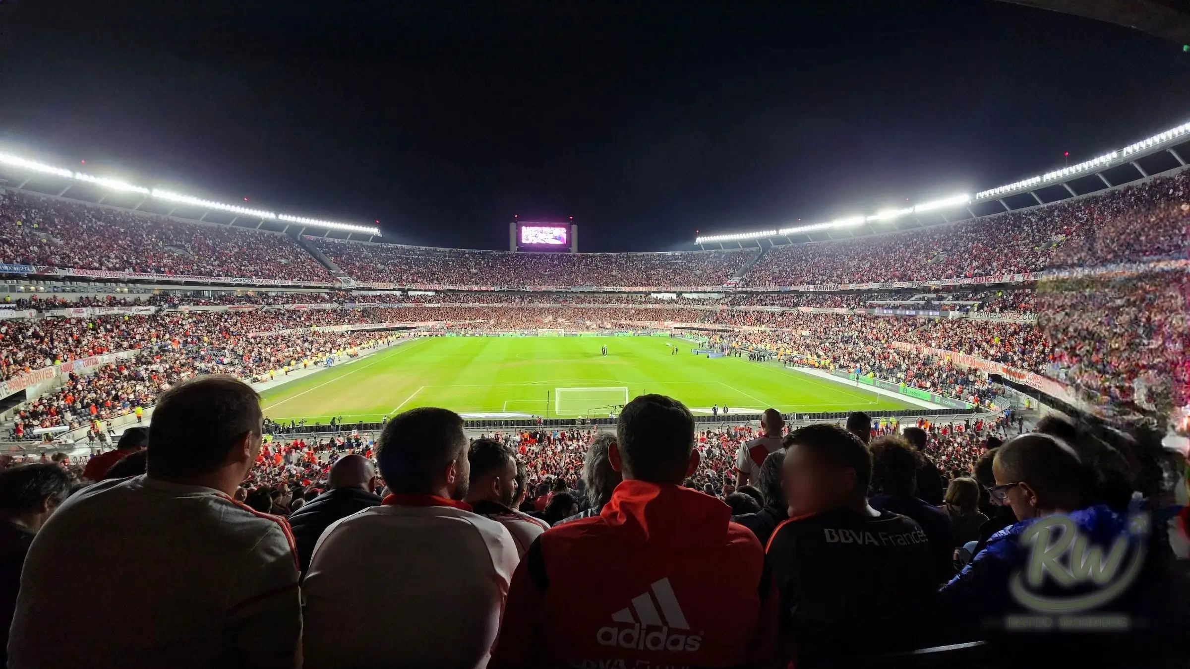 A wide view across the Estadio Monumental bowl at night, supporters in the foreground and the lit pitch below