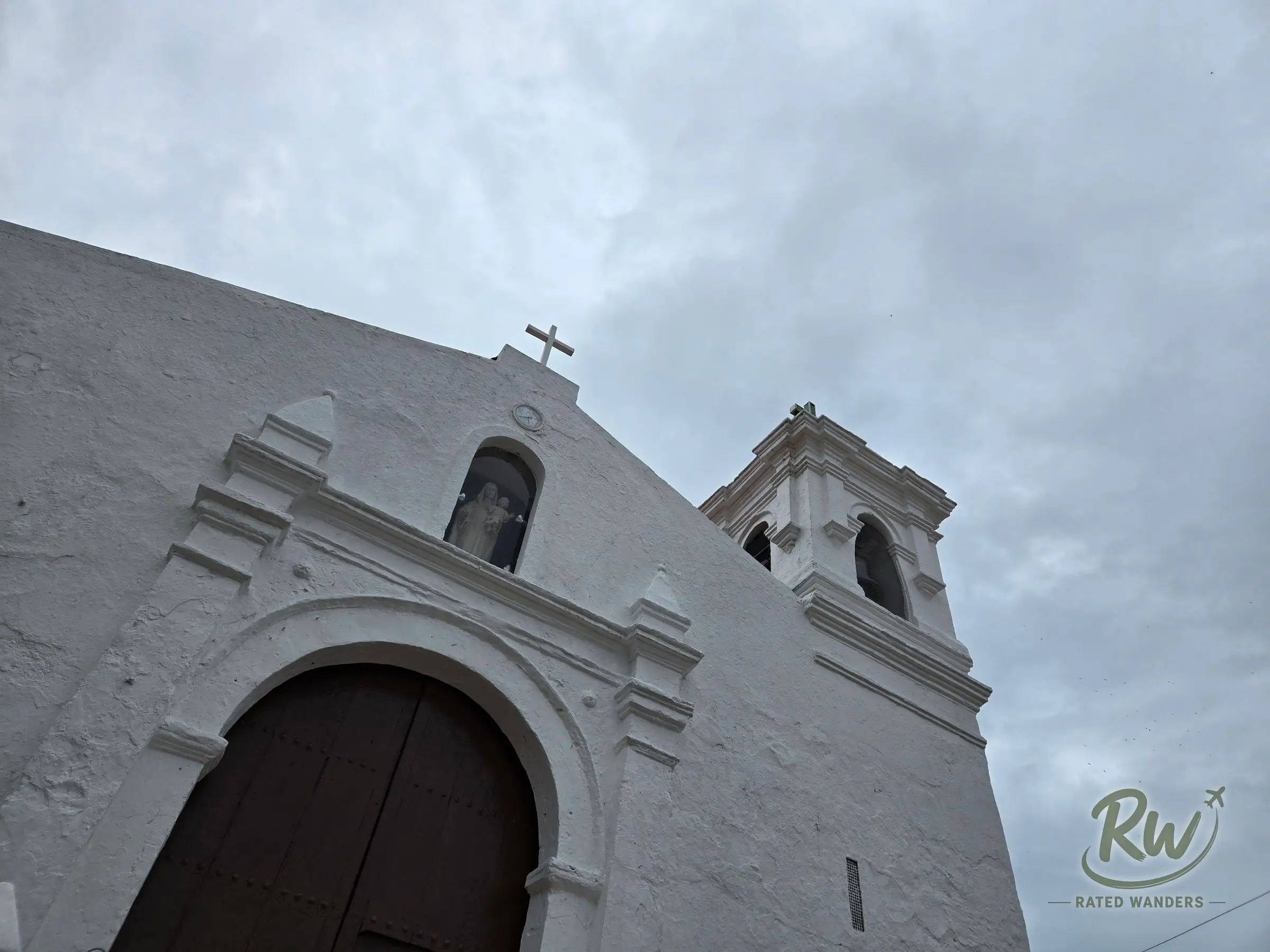 The whitewashed facade and bell tower of Parroquia San Pedro de Taboga seen from below against a grey sky