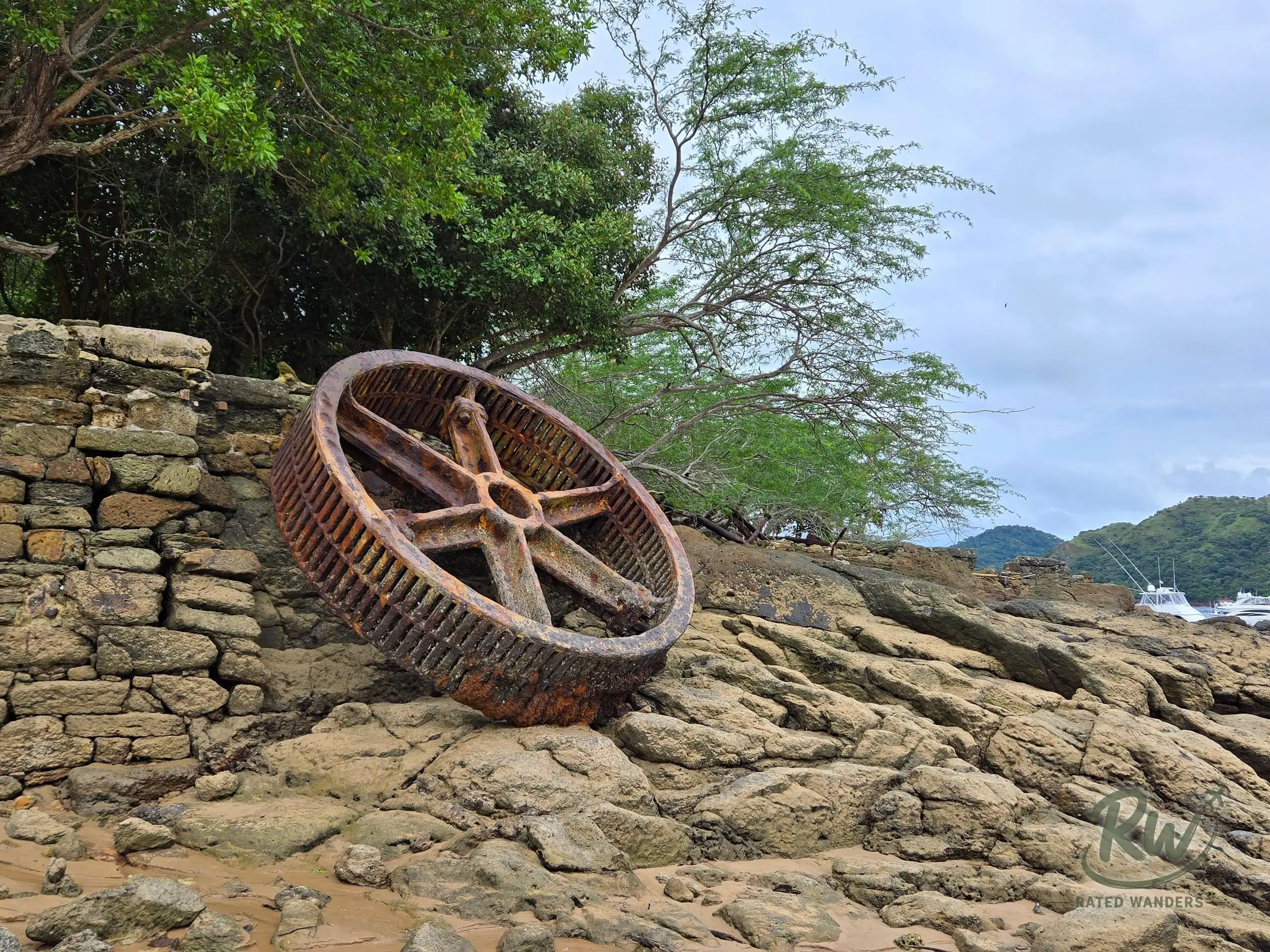 A large rusted iron wheel leaning against an old stone wall on the rocks at the water’s edge