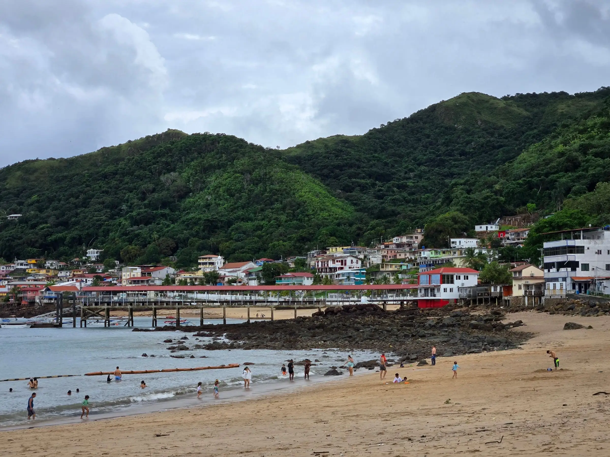 Taboga Island’s main beach with swimmers in the shallows, the village pier behind them and forested hills above