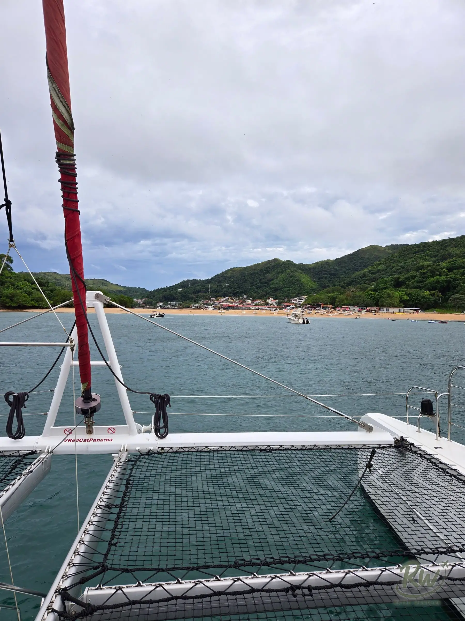 The bow of a catamaran with its trampoline nets and furled red sail, the island’s beach ahead across the water
